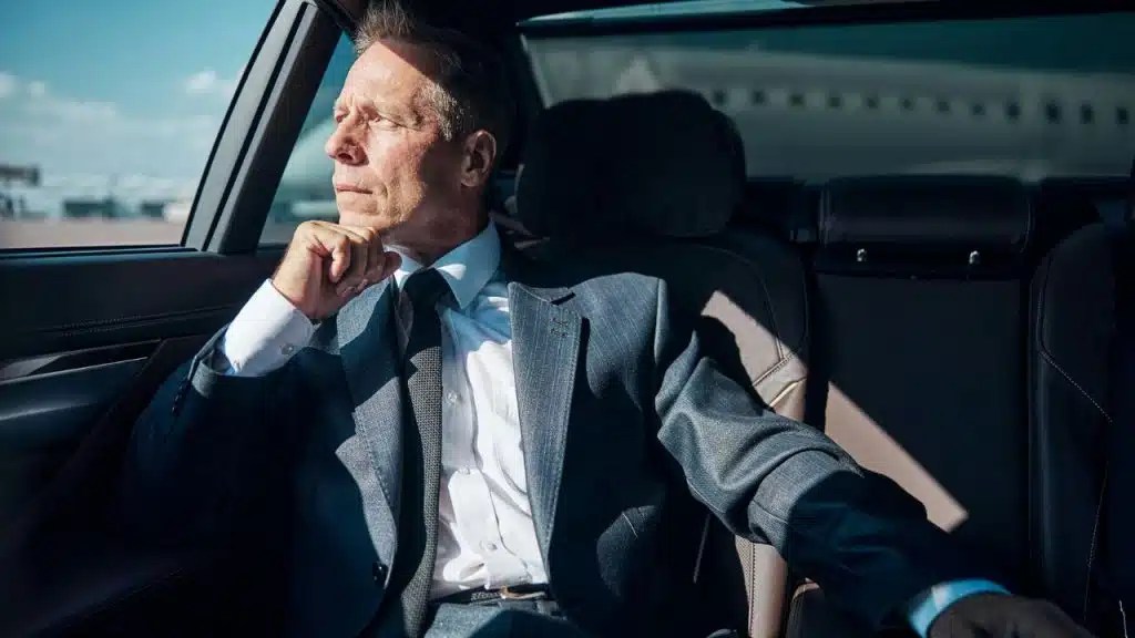 A businessman in a suit is sitting in the back of a car, looking out the window.