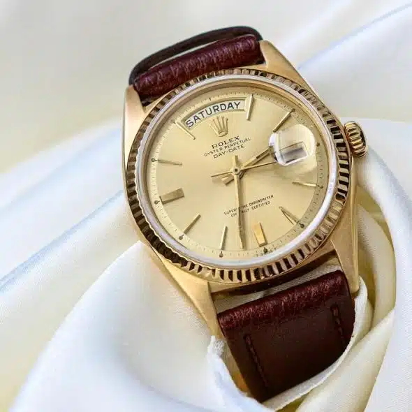 An elegant gold Rolex Day-Date watch with a champagne dial and brown leather strap, resting on white satin fabric.