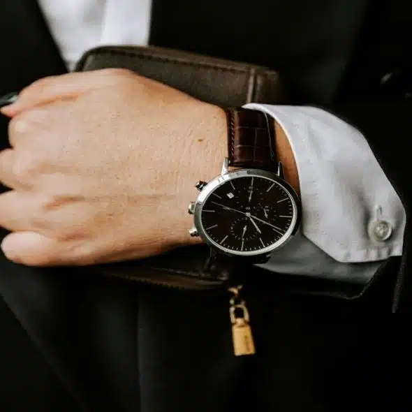 A suited hand wearing a brown leather watch holding a wallet.