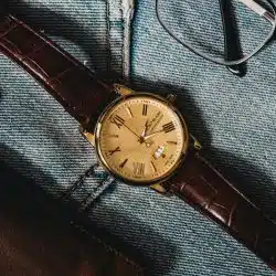 A gold wristwatch with a brown leather strap resting on denim next to eyeglasses.