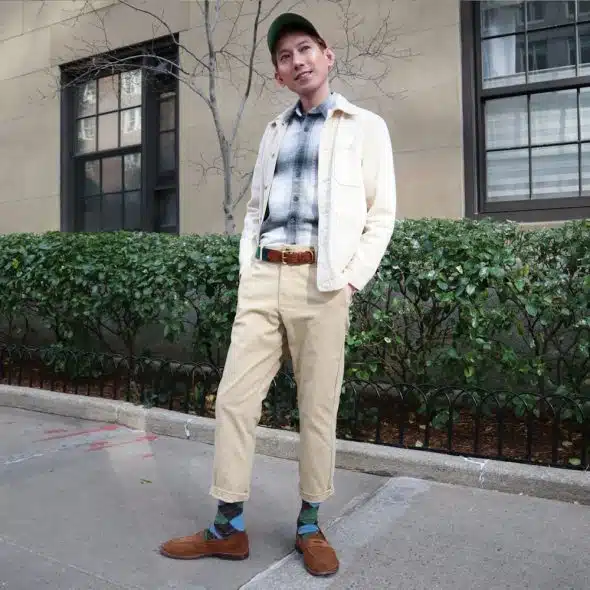 A man with a green cap standing outside wearing a white jacket, a plaid shirt, khaki pants, and brown shoes.