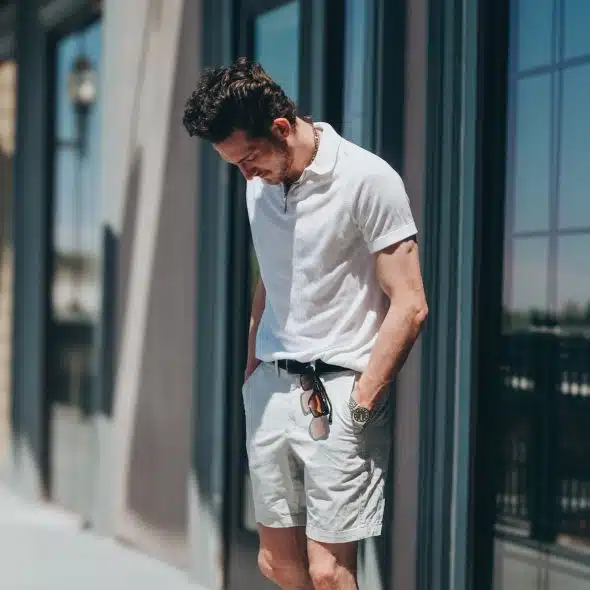 A man with dark, curly hair looking down, wearing a white polo shirt, white shorts, a black belt, and sunglasses tucked into his shirt.