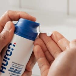A close-up of hands dispensing Huron Broad Spectrum SPF 30 Face Moisturizer from a white and blue pump bottle into a palm.