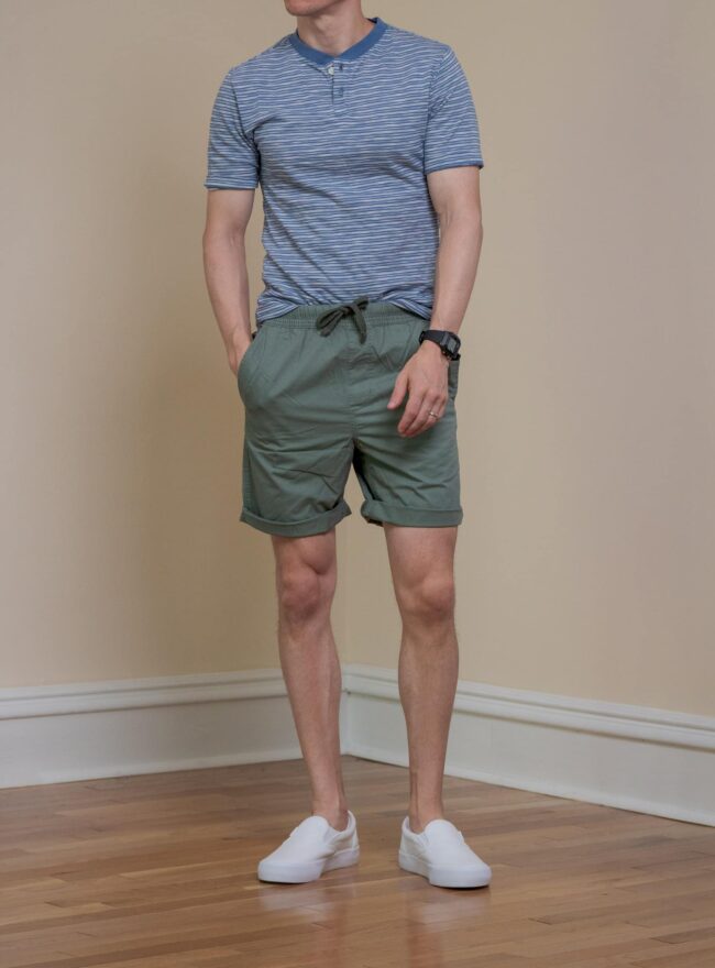 Man wearing a blue horizontally striped t-shirt partially tucked into green shorts, paired with white sneakers worn without socks.