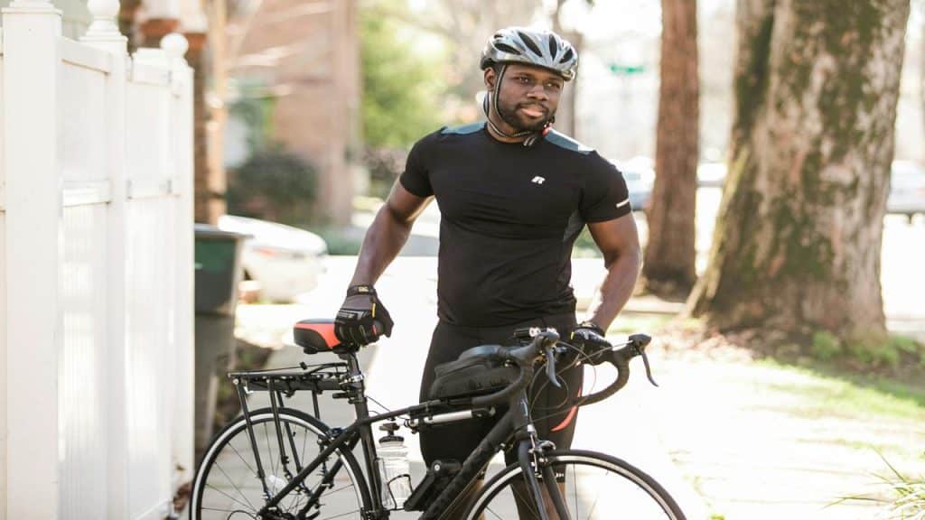 A man in cycling gear standing outdoors with his road bike, wearing a helmet and gloves on a sunny day.