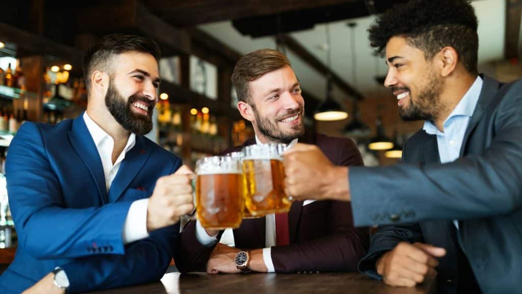 Three men in suits are toasting with beer mugs in a bar.