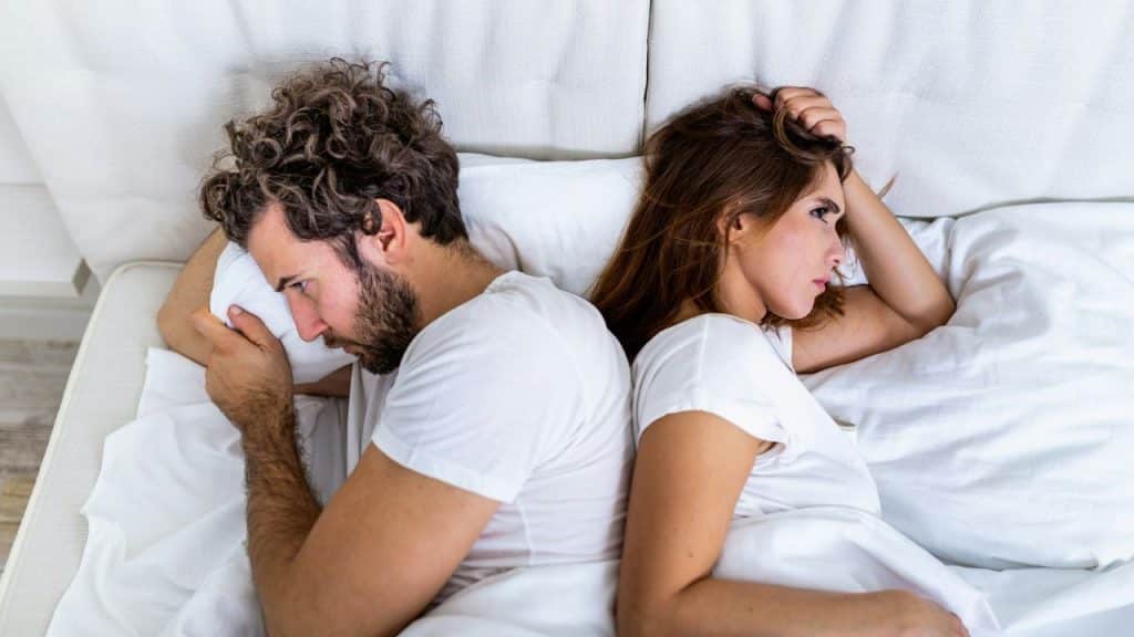 A couple lies in bed back to back, looking upset and distant.