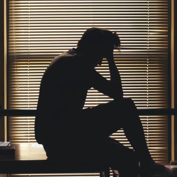 A silhouetted person sitting by a window with blinds, head in hand.