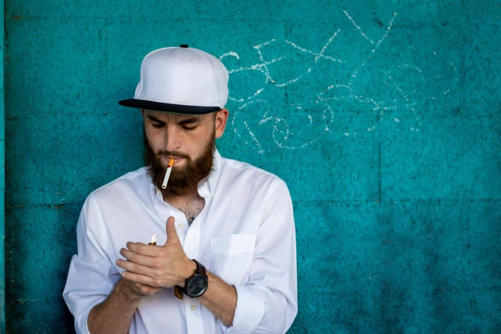 The bearded man is lighting a cigarette.