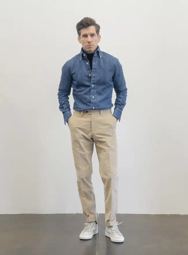 A man standing against a white background, wearing a denim shirt over a black turtleneck, with beige pants and white sneakers.