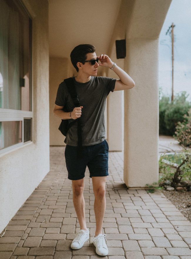 A man standing on a porch wearing a gray t-shirt, navy shorts, white sneakers, and sunglasses.