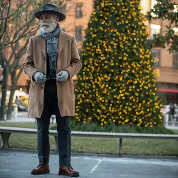 A man with a white beard wearing a brown long coat, a light blue scarf, and gray gloves, holding glasses, standing outdoors with a Christmas tree in the background.